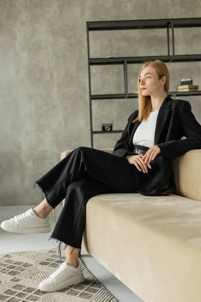 Full length of stylish and pensive woman in black clothes and white sneakers looking away on couch in loft style living room — Stock Photo