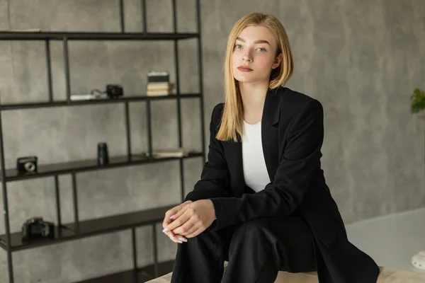 Young blonde woman in black blazer and pants sitting in modern apartment and looking at camera — Stock Photo