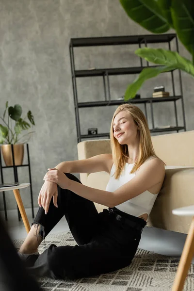 Happy blonde woman in black jeans sitting on floor near sofa and smiling with closed eyes — Stock Photo