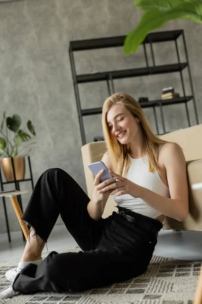 Smiling blonde woman with smartphone sitting on rug near couch in modern living room — Stock Photo