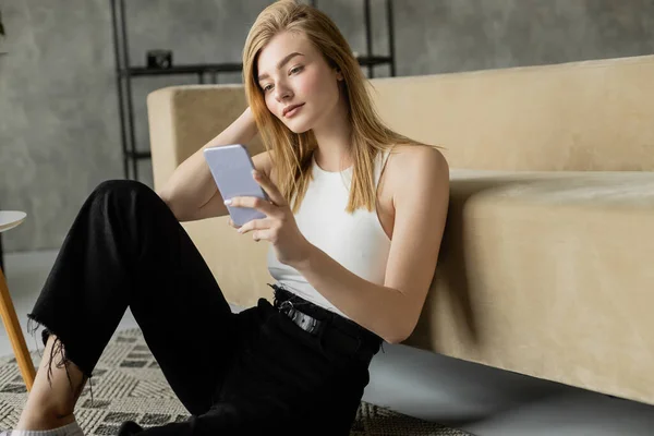 Blonde woman in white tank top and black pants using mobile phone on floor near sofa — Stock Photo