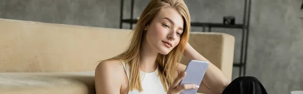 Young blonde woman with mobile phone sitting near modern couch at home, banner — Stock Photo
