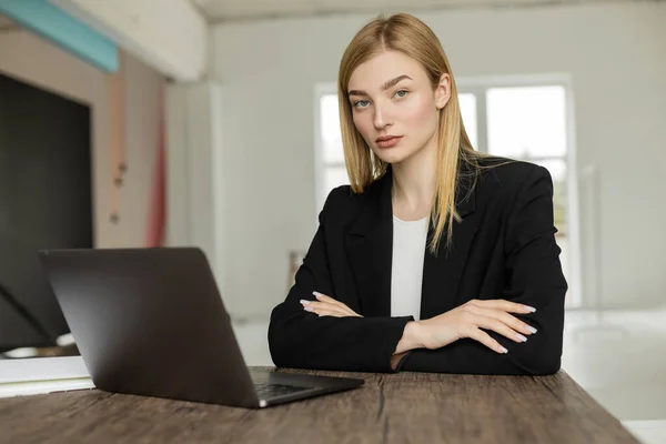 Young and serious coach looking at camera while sitting with crossed arms near laptop — Stock Photo