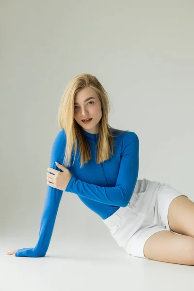 Stylish blonde woman in blue turtleneck and white shorts smiling at camera while sitting on grey — Stock Photo