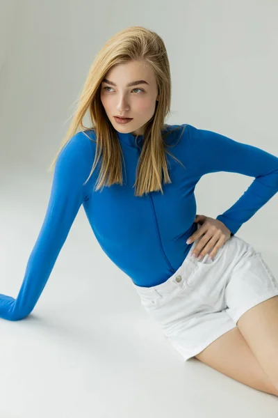 Blondine in blauem Langarmshirt und weißen Shorts schaut weg, während sie mit der Hand an der Hüfte auf grauem Hintergrund posiert — Stockfoto