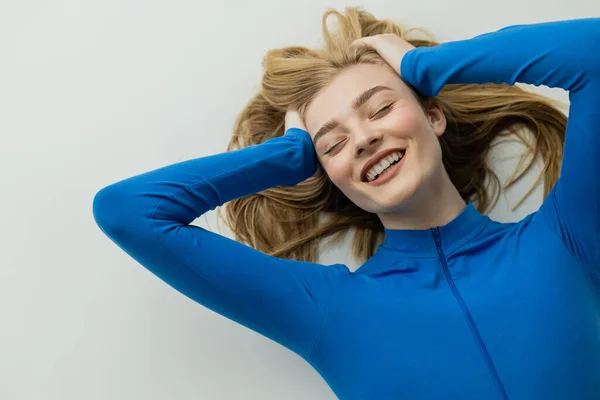 Top view of excited woman with closed eyes touching hair and laughing while lying on grey background — Stock Photo
