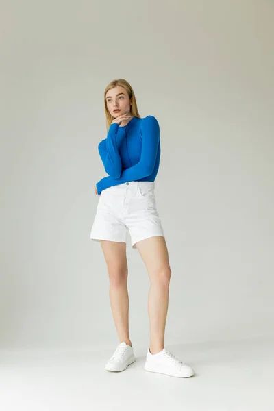 Longitud completa de la esbelta mujer en camisa azul de manga larga y pantalones cortos blancos con zapatillas de deporte mirando hacia otro lado sobre fondo gris - foto de stock
