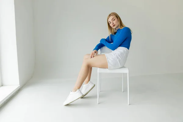 Femme élégante en chemise bleue à manches longues et short blanc assis sur la chaise et regardant la caméra sur fond gris — Photo de stock