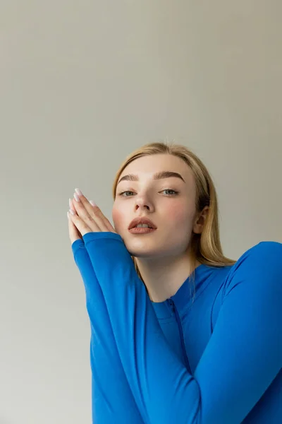 Blonde woman in blue long sleeve shirt holding hands near face and looking at camera isolated on grey — Stock Photo