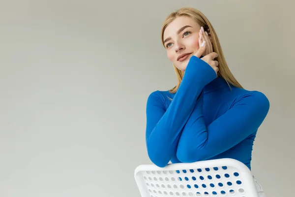 Smiling and dreamy woman in bright blue turtleneck holding hands near face while sitting on chair and looking away isolated on grey — Stock Photo