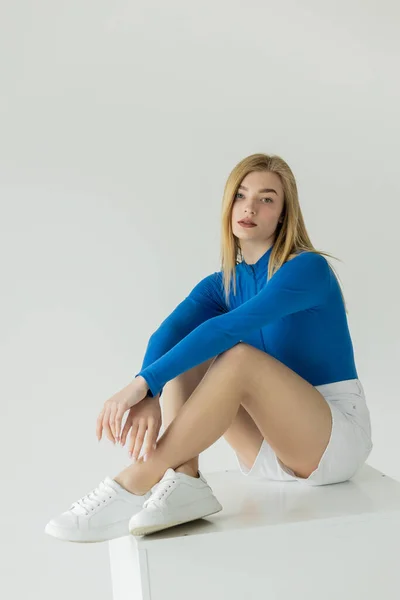 Jolie femme en chemise bleue à manches longues et baskets assises sur cube blanc et regardant la caméra isolée sur gris — Photo de stock