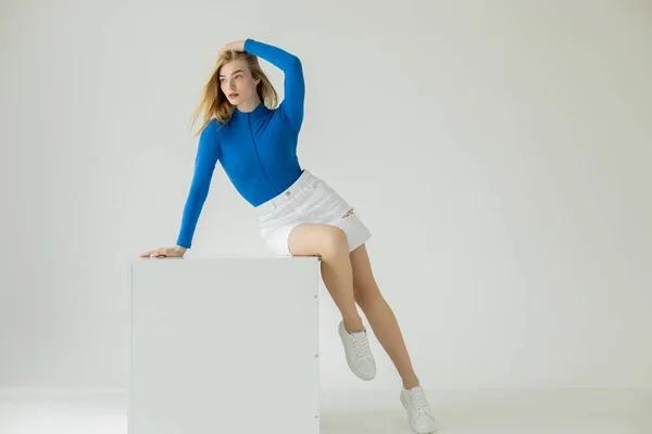 Full length of blonde woman in white sneakers and blue turtleneck sitting on cube and looking away on grey background — Stock Photo