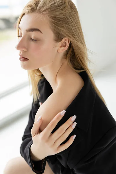 Blonde woman in black blazer touching bare shoulder while posing with closed eyes at home — Stock Photo