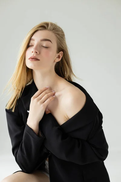 Mulher loira sexy em blazer preto posando com ombro nu e olhos fechados isolados em cinza — Fotografia de Stock