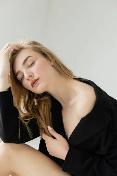 Seductive woman with bare shoulder posing in black blazer with closed eyes isolated on grey — Stock Photo