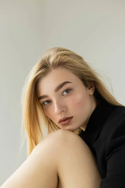 Portrait de femme blonde en veste noire appuyée sur le genou nu tout en regardant la caméra isolée sur gris — Photo de stock