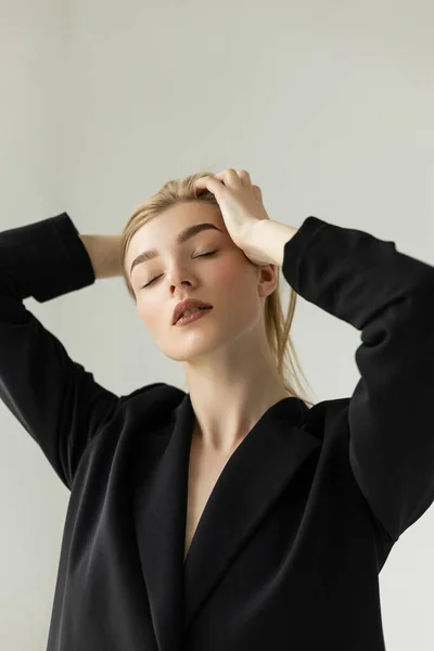 Sensual young woman in black blazer touching blonde hair while posing with closed eyes isolated on grey — Stock Photo
