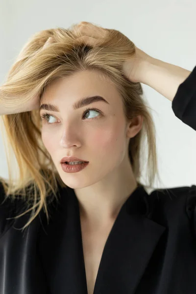 Portrait of pretty woman in black jacket adjusting blonde hair and looking away isolated on grey — Stock Photo