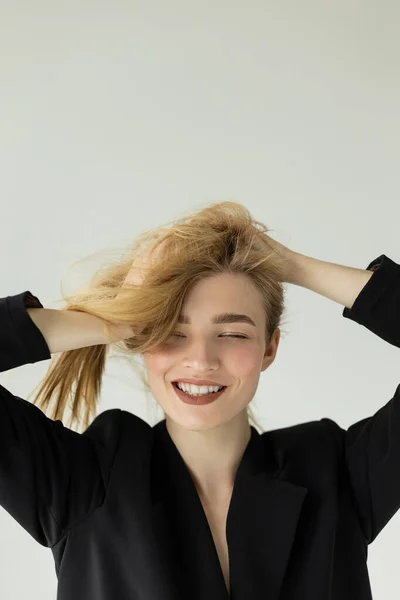 Joyful woman in black jacket touching blonde hair and smiling with closed eyes isolated on grey — Stock Photo