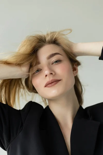 Portrait of positive blonde woman with natural makeup adjusting hair and smiling at camera isolated on grey — Stock Photo