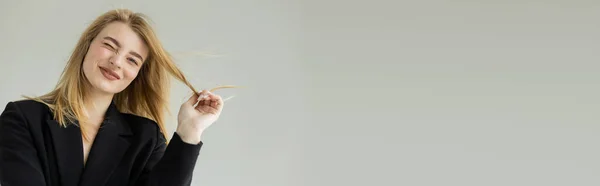 Fröhliche blonde Frau in schwarzem Sakko mit Haaren und zwinkerndem Blick in die Kamera vereinzelt auf grau, Banner — Stockfoto