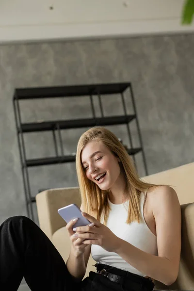 Young excited woman looking at smartphone and laughing near couch in living room — Stock Photo