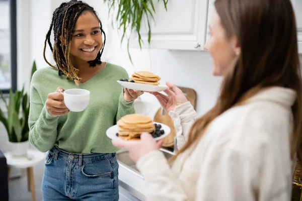 Молодая женщина держит тарелки с вкусными блинами около счастливой африканской американской подруги с чашкой — стоковое фото
