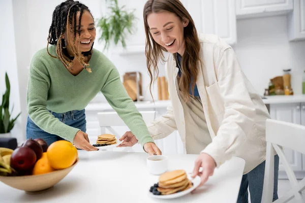 Веселая лесбиянка, подающая тарелки с вкусными блинчиками рядом с африканской американской подружкой — стоковое фото