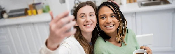 Alegre lesbiana mujer tomando selfie en smartphone con alegre africana americana novia, bandera - foto de stock