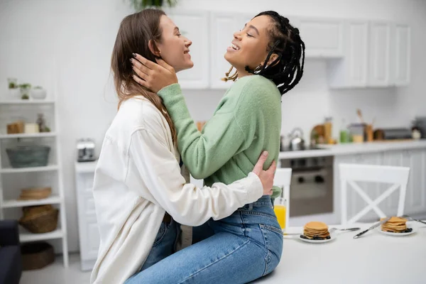 Joyful and lesbian african american woman sitting on dining table and hugging with happy girlfriend — Stock Photo