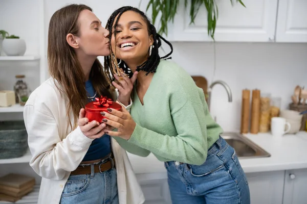 Щаслива лесбіянка цілує щоку задоволеної афроамериканської дівчини, що тримає червону подарункову коробку в день валентинки — стокове фото
