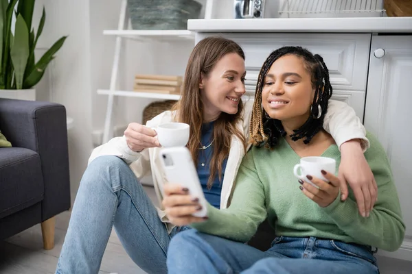 Lesbienne afro-américaine femme tenant smartphone près de petite amie heureuse avec tasse tout en étant assis sur le sol de la cuisine — Photo de stock