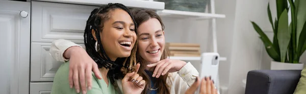Изумленная американская лесбиянка из Африки, использующая смартфон рядом с улыбающейся девушкой, баннер — стоковое фото