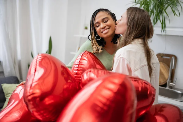 Glücklich lesbische Frau küsst die Wange ihrer afrikanisch-amerikanischen Freundin am Valentinstag in der Nähe roter Luftballons — Stockfoto