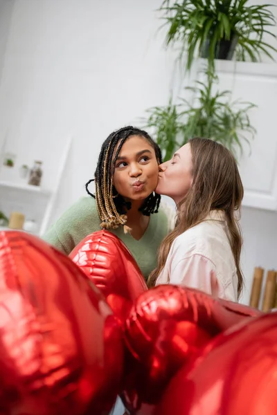 Young lesbian woman kissing cheek of african american girlfriend near red balloons on valentines day — Stock Photo