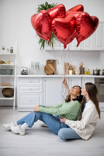 Felice interrazziale e lesbiche donne sedute nel pavimento e guardando palloncini rossi il giorno di San Valentino — Foto stock