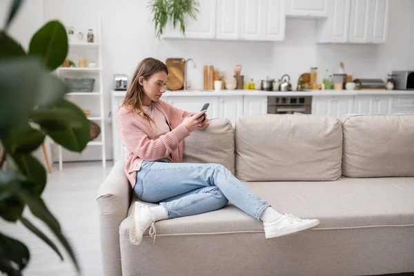 Junge Frau sendet Nachrichten auf dem Smartphone, während sie auf der Couch liegt — Stockfoto