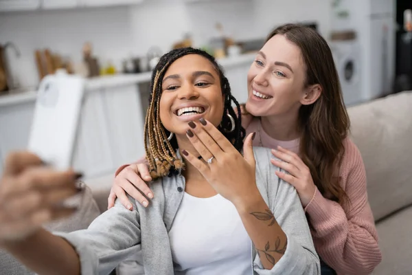 Счастливая африканская американка и лесбиянка, показывающая обручальное кольцо, делая селфи с улыбающейся девушкой — стоковое фото