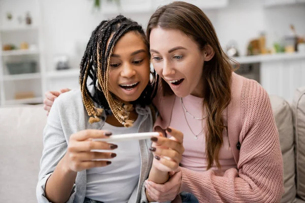 Amazed multiethnic lesbian couple looking at pregnancy test with positive result — Stock Photo