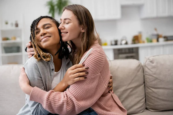 Glücklich afrikanisch-amerikanische lesbische Frau hält Schwangerschaftstest und Umarmung mit fröhlicher Freundin — Stockfoto