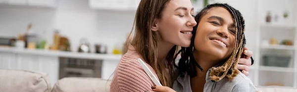 Happy african american and lesbian woman holding pregnancy test and hugging with cheerful girlfriend, banner — Stock Photo