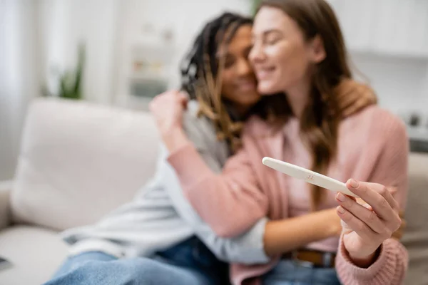 Feliz africano americano y lesbiana mujer abrazando novia con embarazo prueba en borrosa fondo - foto de stock