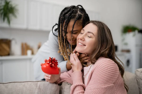 Африканская американская лесбиянка обнимает радостную девушку с подарком в форме красного сердца — стоковое фото
