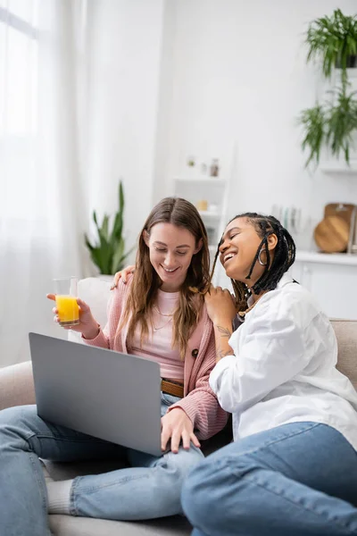 Fröhliche afrikanisch-amerikanische lesbische Frau stützt sich auf glückliche Freundin mit Laptop, während sie von zu Hause aus arbeitet — Stockfoto