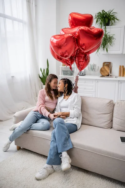 Cheerful lesbian woman holding heart-shaped balloons and sitting on couch with african american girlfriend — Stock Photo
