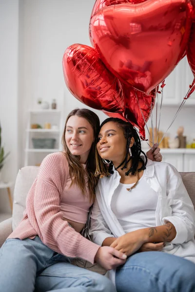 Mujer lesbiana feliz sosteniendo globos en forma de corazón y sentado en el sofá con la novia afroamericana tatuada - foto de stock