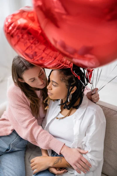 Vista aerea di donna lesbica con palloncini rossi e seduta sul divano con fidanzata afroamericana tatuata — Foto stock
