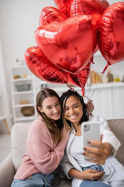 Positive and multiethnic lesbian couple taking selfie near red balloons on valentines day — Stock Photo