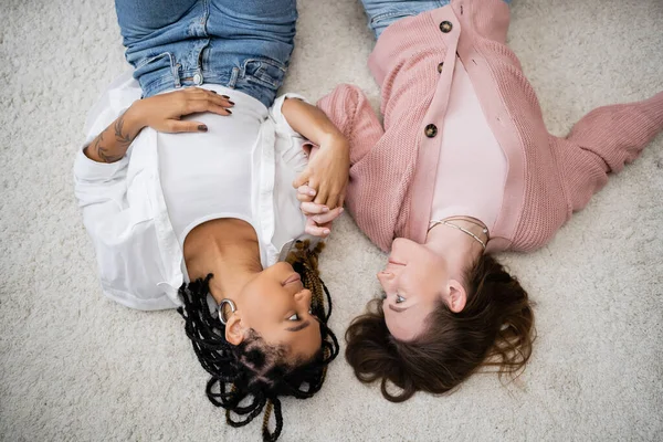 Top view of positive and multiethnic lesbian couple lying on soft carpet and holding hands — Stock Photo