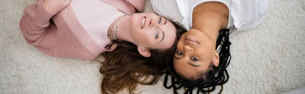 Top view of positive and multiethnic lesbian couple lying on soft carpet, banner — Stock Photo